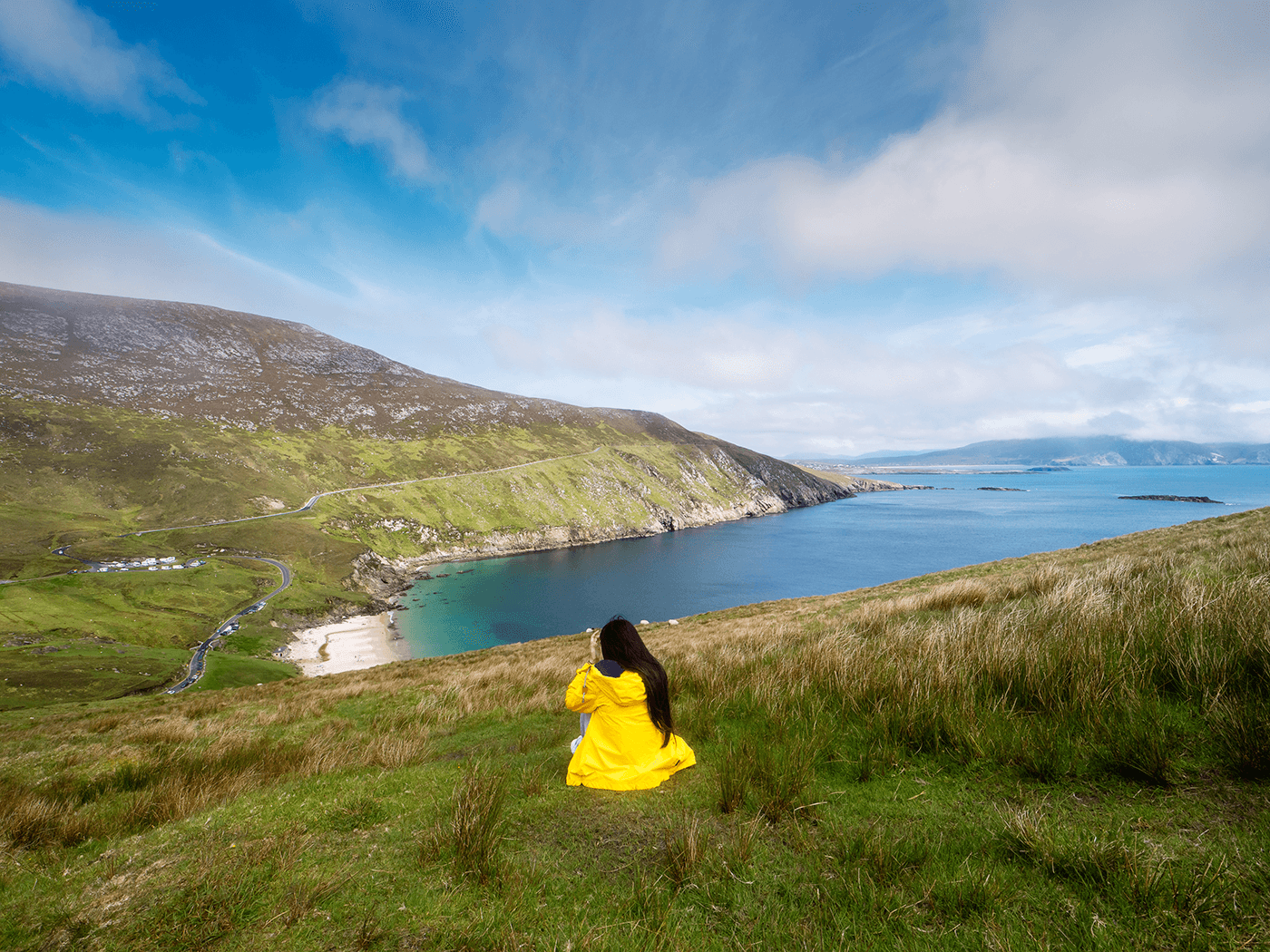 Teenager with long dark brown hair in a yellow jacket looks out over Keem Bay in Co. Mayo